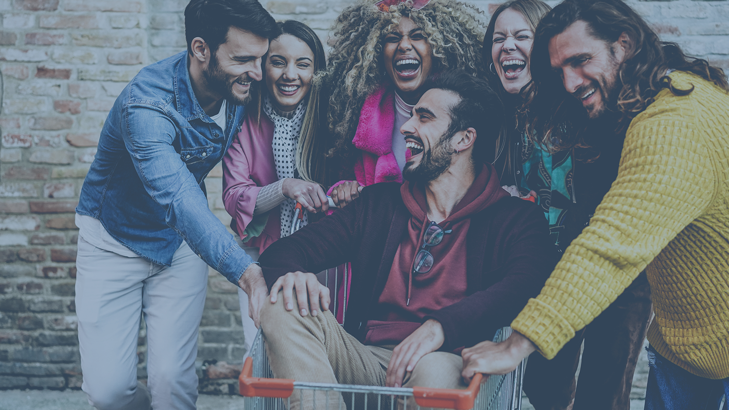 Photo of six young and diverse professionals, fashionably dressed in colorful, business-professional clothing. One is seated in a shopping cart. Others surround the back and sides of the cart and have their hands on the cart. All are laughing.