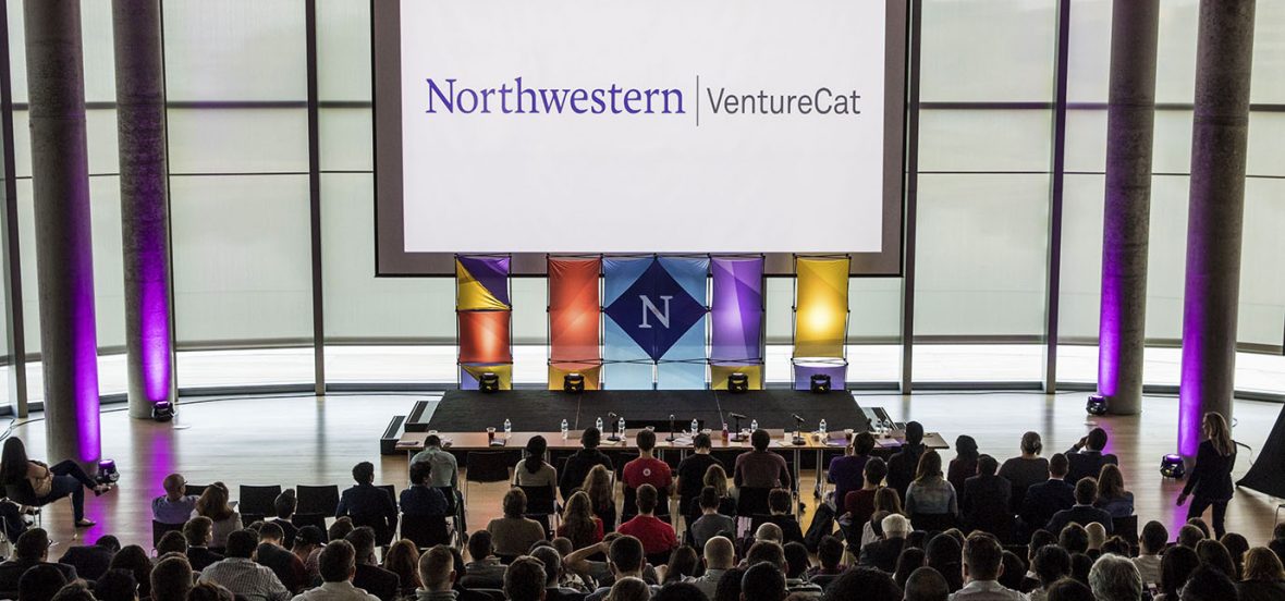 Photo of the VentureCat stage in the White Auditorium of Kellogg Global Hub. A large projection screen hangs from the ceiling and displays the Northwestern Wordmark along with the VentureCat title. A banner with the Northwestern N in a purple diamond hangs from the center of the projection screen. Two colorful banners flank each side of the logo banner. A full crowd is seated in the lower part of the photo.