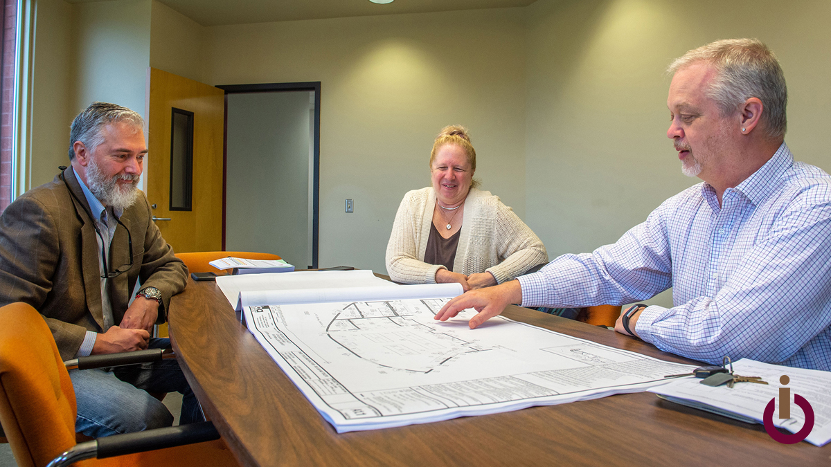 Three people sitting at a conference table, looking at architectural plans, and discussing. A small iFERM logo is in the lower-right corner of the photo.