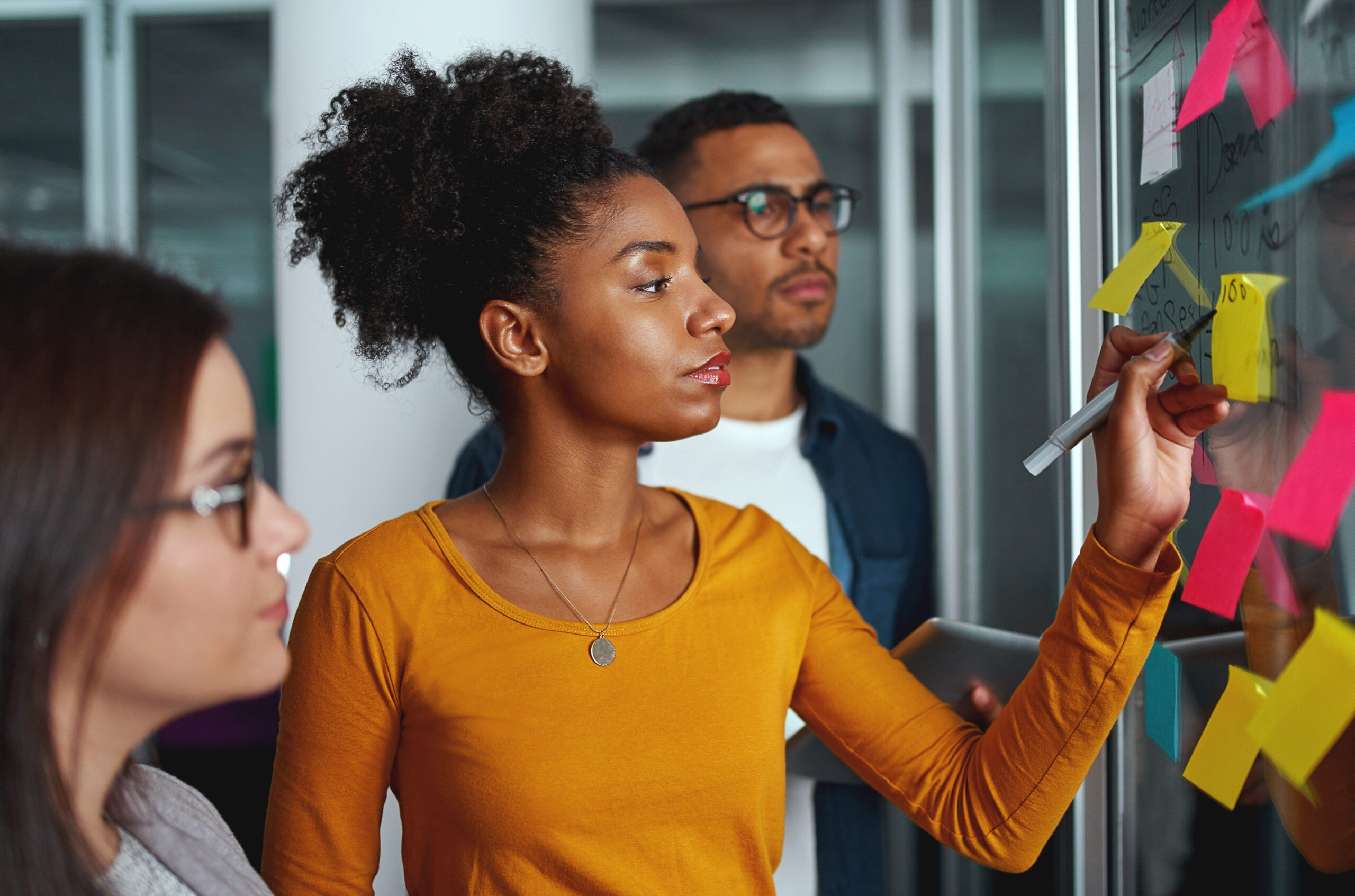 Young creative businesswoman standing with her colleagues writing new ideas on sticky notes over glass wall. The three represent minority or underrepresented populations in technology entrepreneurship.